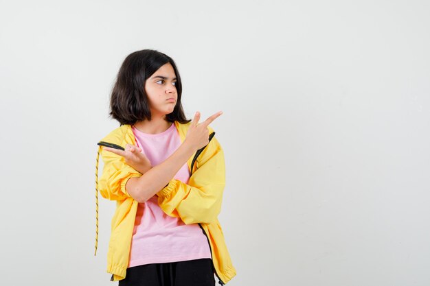 Teen girl in yellow tracksuit, t-shirt pointing aside with crossed arms and looking thoughtful , front view.