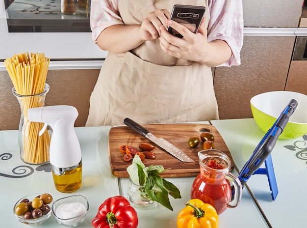 Foto adolescente prepara un tutorial virtuale online sul libro di testo e guarda la ricetta digitale sul telefono cellulare touch screen mentre prepara cibo sano in cucina a casa