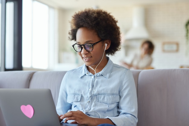 Teen schoolgirl wearing earphones using laptop having online lesson sitting on the couch at home