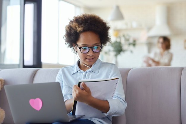 Teen schoolgirl wearing earphones using laptop looking at her notebook sitting on the couch at home