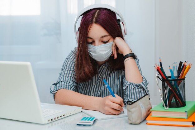 ウイルスに対して医療マスクを持つ十代の女子高生は、宿題をしている家のラップトップを勉強して床のリビングルームのソファの学生を座っています。