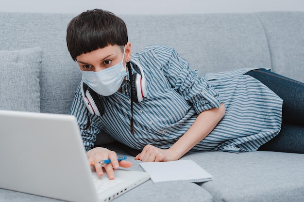 Teen schoolgirl with medical mask against viruses sitting floor living room sofa student studying home laptop doing homework