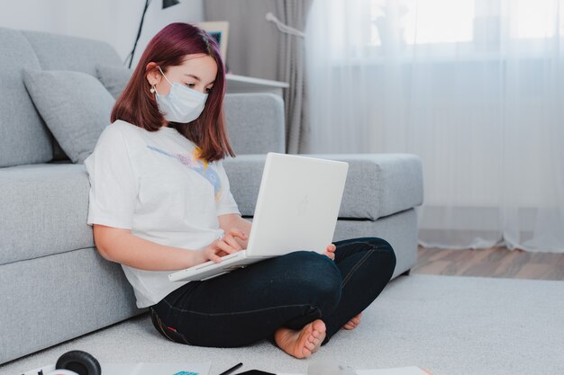 Teen schoolgirl with medical mask against viruses sitting floor living room sofa student studying