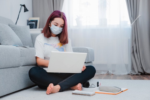 Teen schoolgirl with medical mask against viruses sitting floor living room sofa student studying