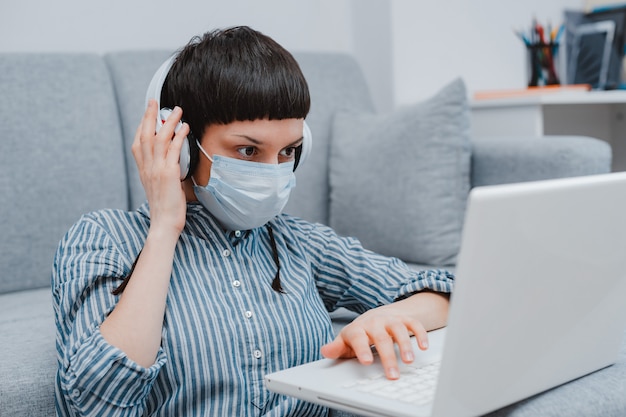 Teen woman with medical mask against viruses sitting floor living room sofa student studying home laptop doing homework