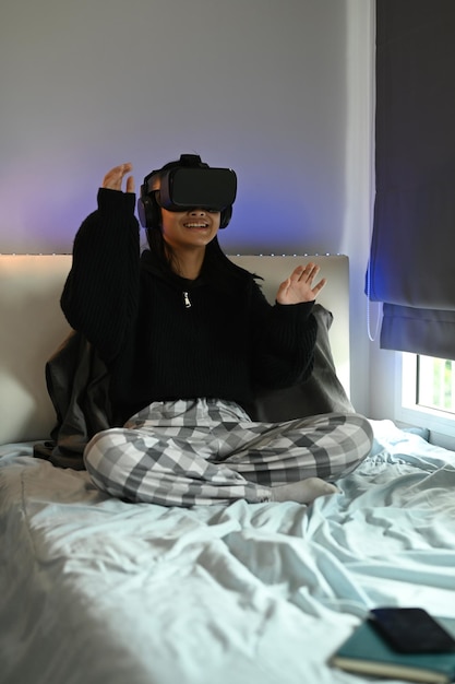 Teenage asian girl experiencing opportunities of gaming in virtual reality headset while sitting in bedroom