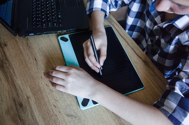 A teenage boy in checked shirt in the process of home training draws using a graphics tablet, which lies on a wooden table.