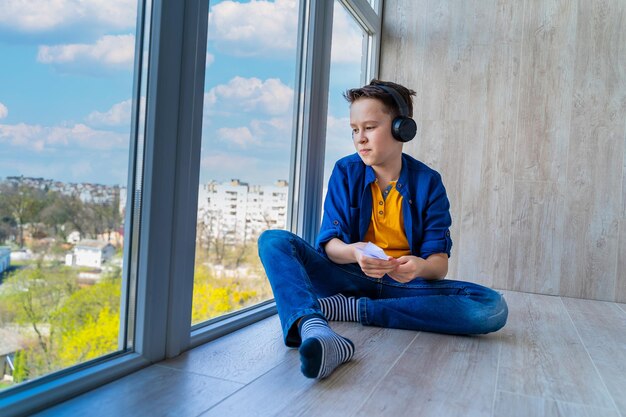 Teenage boy in headphones sitting at floor Boy holding paper airplane in hands looking at city through the window