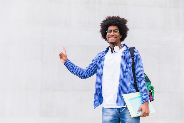 Photo teenage boy holding books in hand pointing his finger upward against white concrete wall