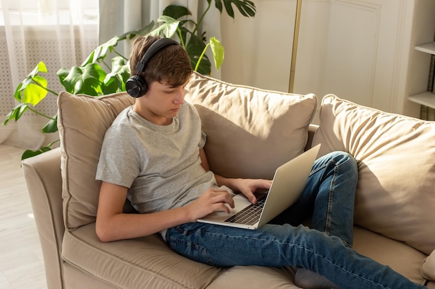 A teenage boy lying on the couch in a sunny room wearing black\
headphones playing on a laptop