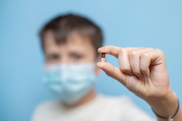 Teenage boy in protective medical mask holds capsule with medication in hands.