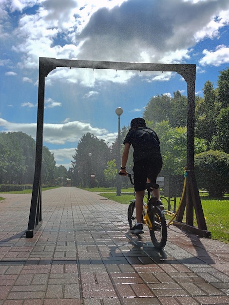 A teenage boy rides a bicycle under a cooling frame in a city park and laughs