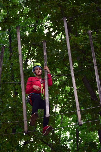 A teenage boy in a safety helmet climbs a hanging ladder in a\
rope amusement park