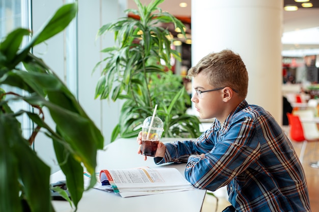 A teenage boy sits at a table in a cafe with a book concept of distance learning college life