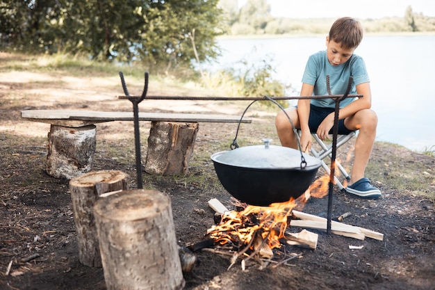 Teenage boy sitting around a campfire in forest in summer Family picnic outdoors Camping life Cooking in cauldron on fire in nature