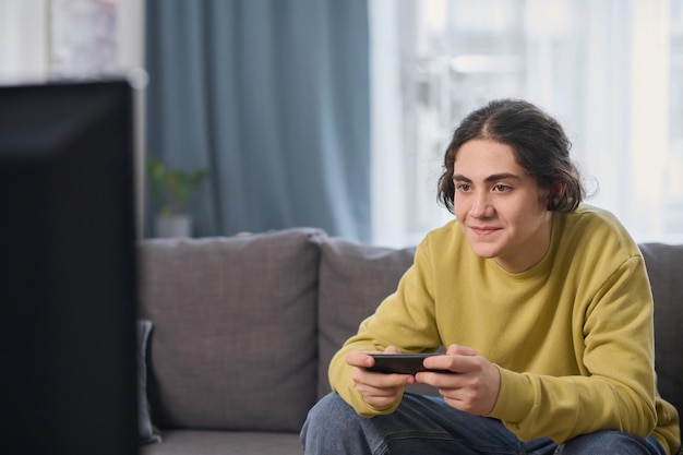 Teenage boy sitting on sofa in front of tv and using joystick to playing video game at home