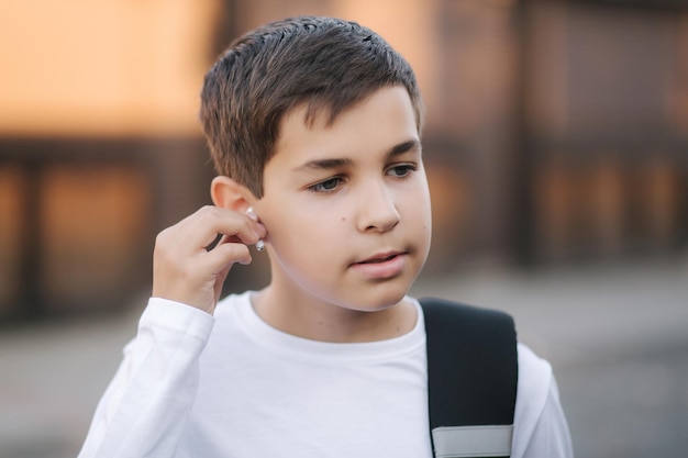 Teenage boy take out a wireless headphones and put then into the ears Young boy in white shirt with backpack Boy listen music