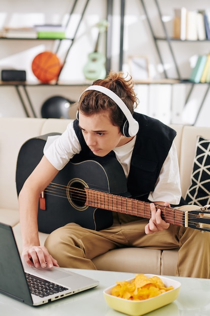 Photo teenage boy in wireless headphones eating potato chips when learning how to play new song on acoustic guitar