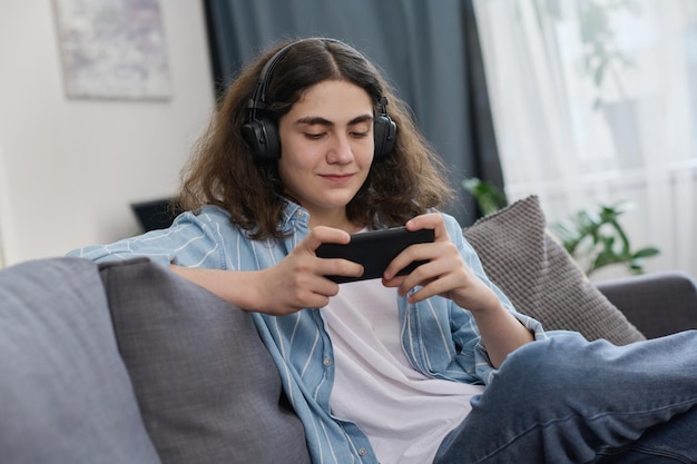 Teenage boy in wireless headphones playing game on smartphone sitting on sofa in the room