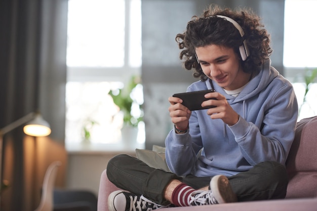 Foto ragazzo adolescente in cuffie wireless seduto sul divano usando il suo telefono cellulare per giocare a giochi online