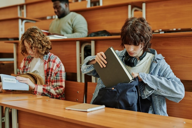 Teenage brunette girl in casualwear putting book into backpack after lesson