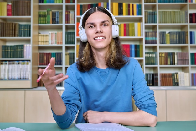 図書館の机に座ってウェブカメラを見ているヘッドフォンの 10 代の大学生 ビデオ通話をする男会議チャット リモートで勉強してオンライン試験を受けるレッスン コース 教育テクノロジー