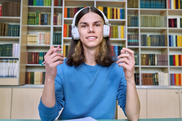 Teenage college student in headphones looking at webcam sitting at desk in library