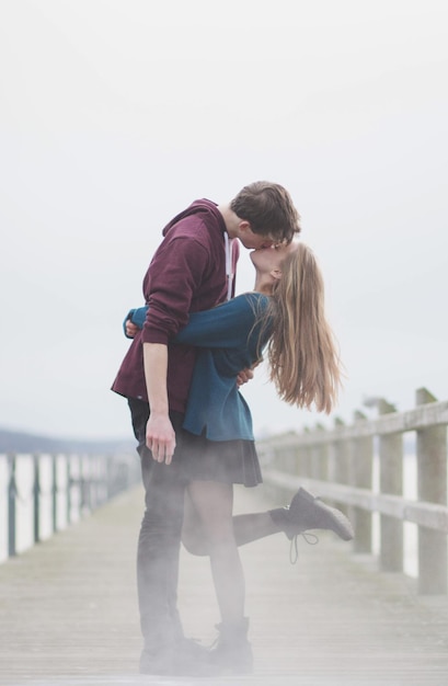 teenage couple kissing while standing on bridge against sky