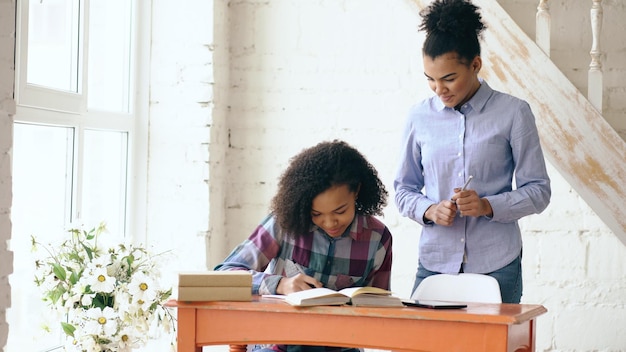 Teenage curly haired young girl sitting at the table learning lessons and her elder sister helps