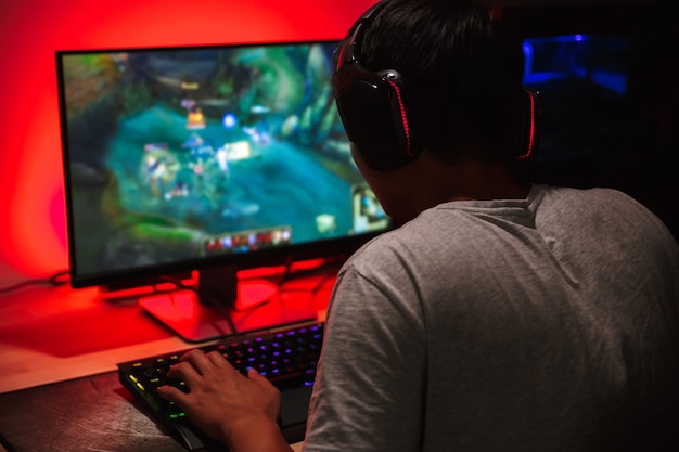 Teenage gamer boy playing video games on computer in dark room, wearing headphones and using backlit colorful keyboard