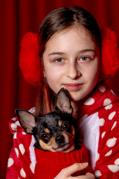 10代の少女とチワワは、赤を背景に自宅で犬用の赤いセーターを着ています。