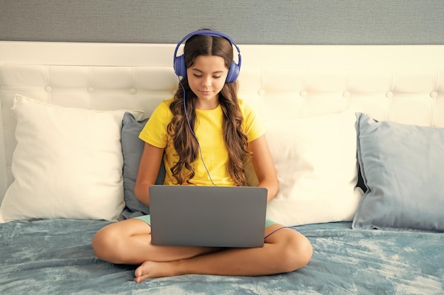 헤드폰을 끼고 있는 10대 소녀 아이는 집에서 침대에서 휴식을 취하며 노트북을 사용하여 음악을 듣습니다. 이어폰을 낀 아이는 컴퓨터에서 인터넷을 검색합니다.