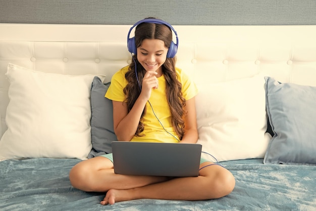 Teenage girl child in headphones relax on bed at home listen to music using laptop Child in earphones browse internet on computer