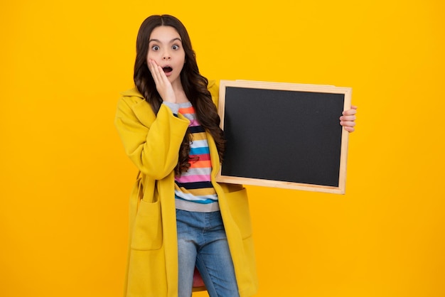 Teenage girl child holding blackboard isolated on a yellow background Shocked amazed face surprised