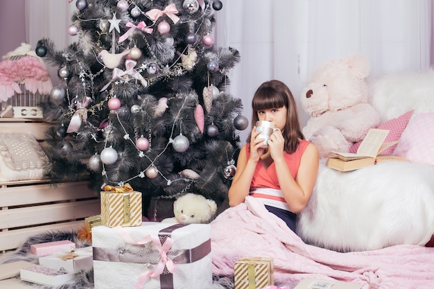 Teenage girl drinks tea in New Year's living room, surrounded by gifts and toys, next to Christmas tree.