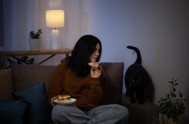Девочка-подросток ест бутерброд с кошкой ночью на диване