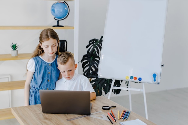 Teenage girl and first grader boy study on laptop together sitting at desk in classroom Educational school process bright room and interesting learning