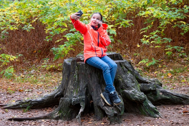 Teenage girl generation z sits on huge tree stump talks on
phone and takes selfie at same time