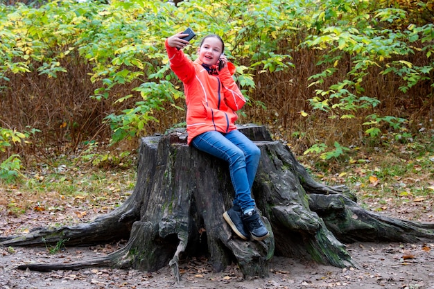 Teenage girl generation z sits on huge tree stump talks on\
phone and takes selfie at same time