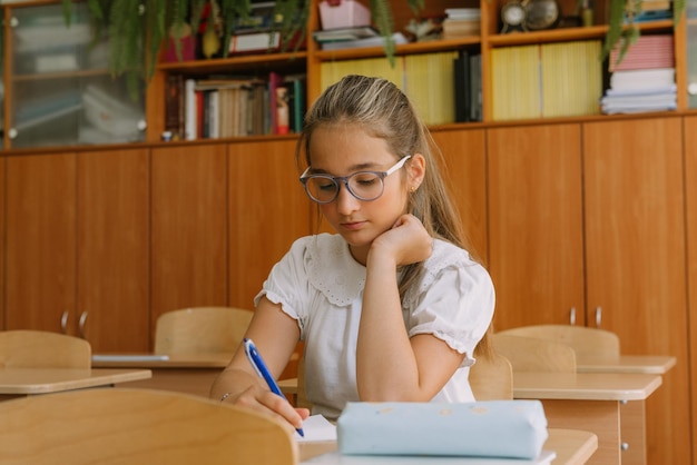 Teenage girl in glasses writing in copybook at desk in classroom