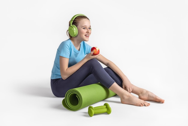 Teenage girl in headphones apple in hand sits on the floor Green dumbbells and exercise mat Rela