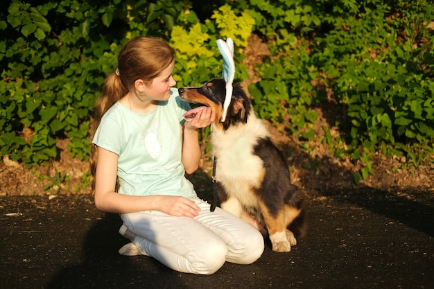 Teenage girl hold funny cute Australian shepherd three colours puppy dog wearing bunny ears. happy Easter. Holiday. Outdoor. In spring park.