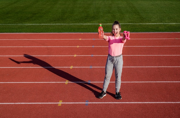 바벨과 물병 휘트니스 코치를 들고 있는 10대 소녀는 학교 체육 수업 아령에서 경기장 체육관 전력 및 근력 아동 훈련에서 워밍업 운동을 준비합니다.