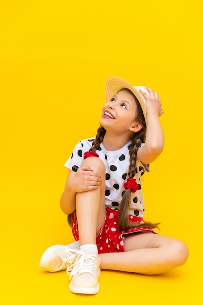 A teenage girl is holding on to a straw hat and sitting in full height A beautiful little lady in shorts is waiting for summer on a yellow isolated background