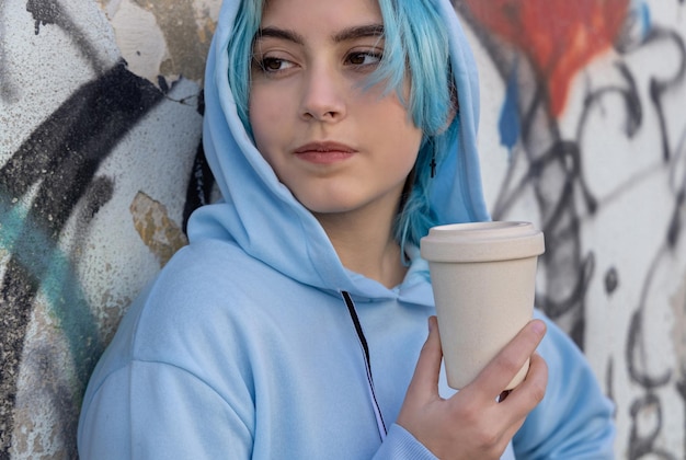 Teenage girl in light blue oversize hoodie with coffee to go and looking aside Blue haired teen girl staying outdoors near graffiti wall Cup mockup Hipster and adolescence concept