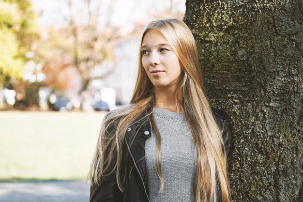 Teenage girl looking away leaning against tree trunk at park