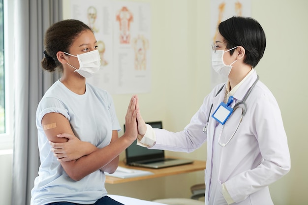 Teenage girl in medical mask giving high five to doctor after getting shot of vaccine against corona...