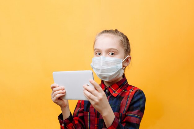 A Teenage Girl In A Medical Mask On A Yellow wall Is Holding A Tablet PC