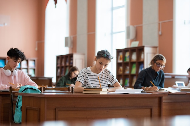 Девочка-подросток и другие учащиеся колледжа сидят за партами в библиотеке и делают записи в тетрадях во время подготовки к семинару