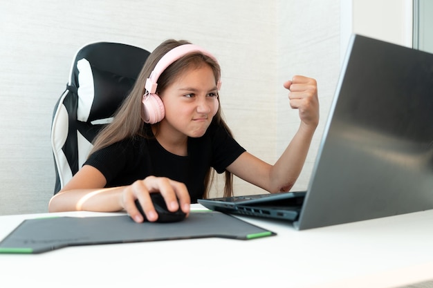 teenage girl in pink headphones is playing computer games on a white background A negative emotion on the face the child shows his fist at the laptop screen